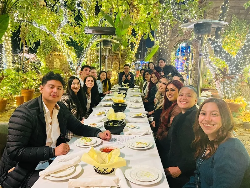 Dental Staff group at table: man in black jacket gestures toward camera; plates, drinks, and warm lighting visible