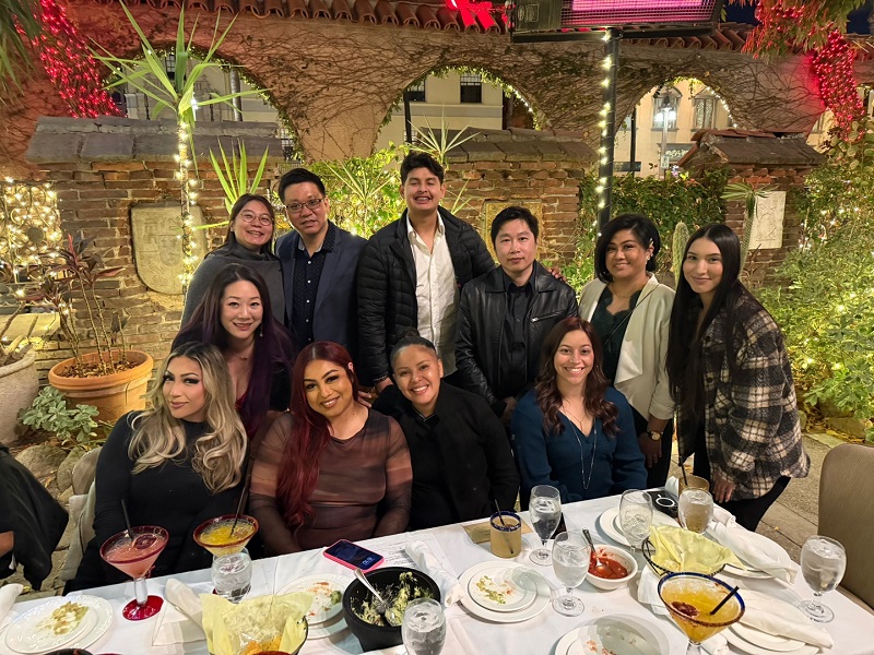 Dental Staff group posing at table; some standing, some seated, with food and drinks, brick wall backdrop