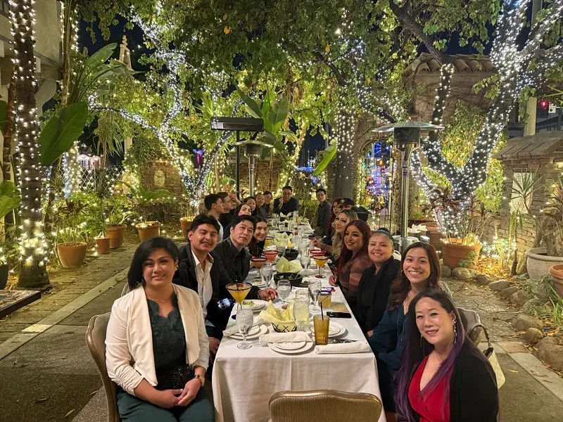 Dental Staff group seated at long outdoor dinner table under string lights and greenery; smiling, festive evening setting